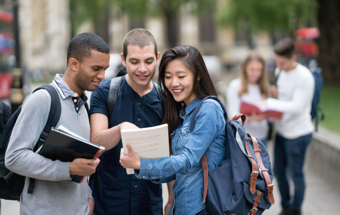 A multi-ethnic group of college or university students look at class materials and smile while outside.
